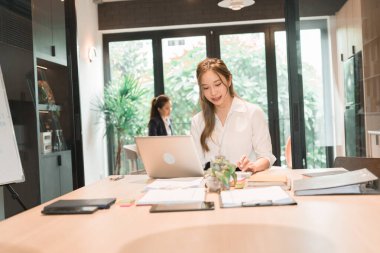 Üç genç iş kadınından oluşan bir grup, belgeler, tablet, dizüstü bilgisayar ve diğer ekipmanlarla birlikte bir masada oturuyorlar. Şirketi geliştirmek için bir tartışma ve planlama oturumu yapıyorlar..