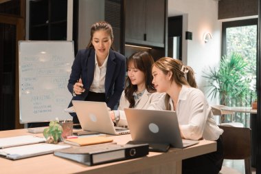 Üç genç iş kadınından oluşan bir grup, belgeler, tablet, dizüstü bilgisayar ve diğer ekipmanlarla birlikte bir masada oturuyorlar. Şirketi geliştirmek için bir tartışma ve planlama oturumu yapıyorlar..