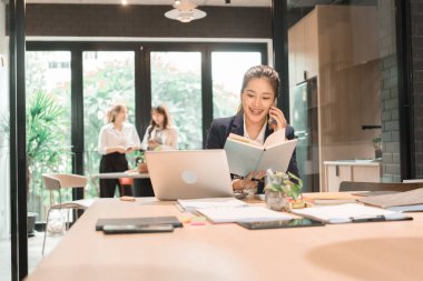 Üç genç iş kadınından oluşan bir grup, belgeler, tablet, dizüstü bilgisayar ve diğer ekipmanlarla birlikte bir masada oturuyorlar. Şirketi geliştirmek için bir tartışma ve planlama oturumu yapıyorlar..