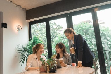 Üç genç iş kadınından oluşan bir grup, belgeler, tablet, dizüstü bilgisayar ve diğer ekipmanlarla birlikte bir masada oturuyorlar. Şirketi geliştirmek için bir tartışma ve planlama oturumu yapıyorlar..
