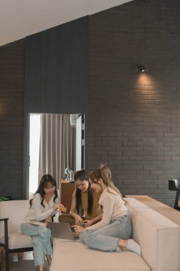 Üç genç Asyalı kadın evde bilgisayar ve kredi kartıyla online alışveriş yapıyor. Rahatlamış ve modern bir oturma odasında birlikte gülümsüyor ve eğleniyorlar..