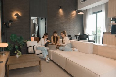 Üç genç Asyalı kadın evde bilgisayar ve kredi kartıyla online alışveriş yapıyor. Rahatlamış ve modern bir oturma odasında birlikte gülümsüyor ve eğleniyorlar..