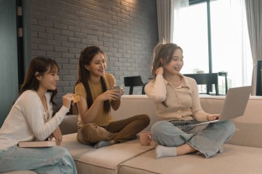 Üç genç Asyalı kadın evde bilgisayar ve kredi kartıyla online alışveriş yapıyor. Rahatlamış ve modern bir oturma odasında birlikte gülümsüyor ve eğleniyorlar..