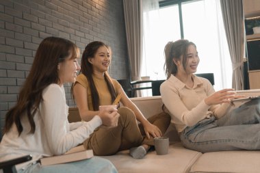 Üç genç Asyalı kadın evde bilgisayar ve kredi kartıyla online alışveriş yapıyor. Rahatlamış ve modern bir oturma odasında birlikte gülümsüyor ve eğleniyorlar..