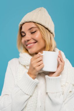 pleased young woman in white sweater and hat holding cup of coffee isolated on blue  clipart