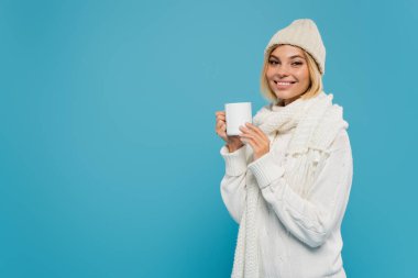 joyful young woman in white sweater and hat holding cup of coffee isolated on blue  clipart