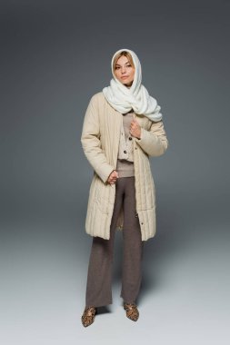 full length of stylish young woman in winter headscarf and coat standing on grey clipart