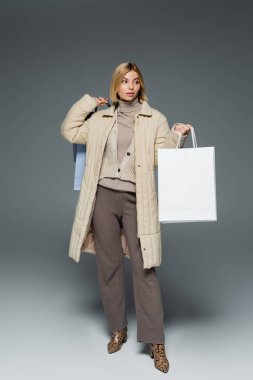 Kışlık ceketli ve balıkçı yakalı moda bir kadın gri arka planda alışveriş torbaları taşıyor. 