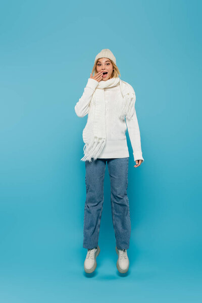 full length of excited young woman in winter outfit covering mouth while levitating on blue 