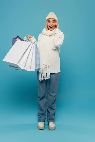 白いセーターと帽子を着た驚くべき女性の完全な長さ青でスマートフォンで話している間にショッピングバッグを保持 — ストック写真