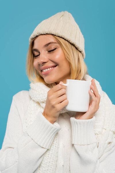 pleased young woman in white sweater and hat holding cup of coffee isolated on blue 