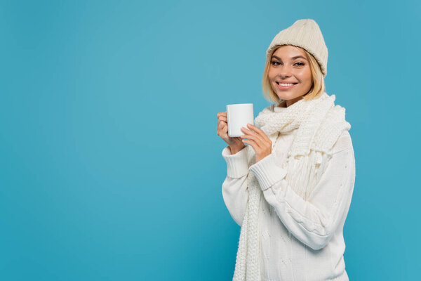 joyful young woman in white sweater and hat holding cup of coffee isolated on blue 