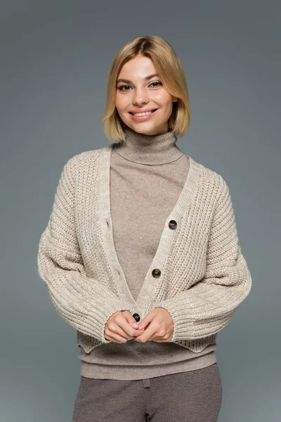 happy blonde woman in turtleneck and cardigan smiling isolated on grey