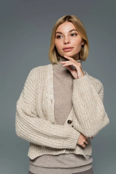 young blonde woman in turtleneck and cardigan looking away isolated on grey