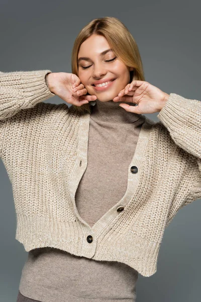 cheerful blonde woman in turtleneck and cardigan smiling with closed eyes isolated on grey