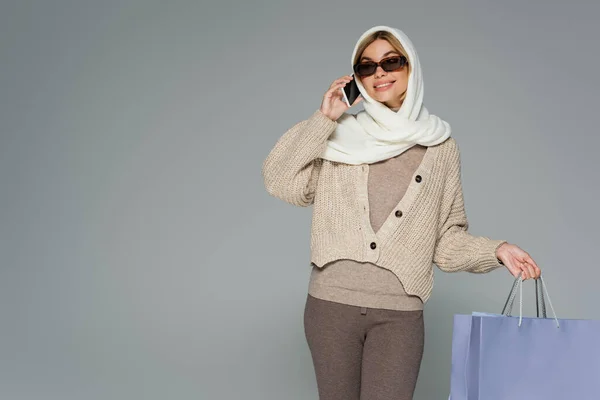 stock image cheerful woman in knitwear and sunglasses holding shopping bags while talking on smartphone isolated on grey