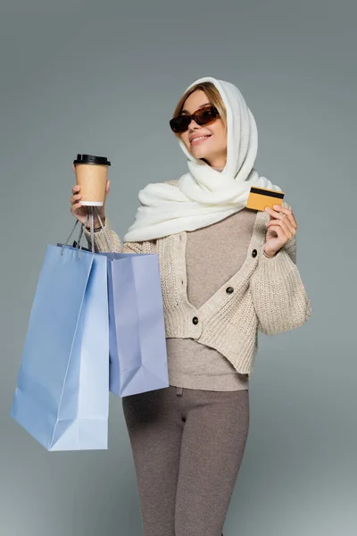 ヘッドスカーフとサングラスをした陽気な女性が紙コップとクレジットカードを持ってグレーに隔離された買い物袋で立っている間 — ストック写真