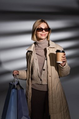 Soyut gri arka planda alışveriş torbaları ve kahve taşıyan kışlık ceketli pozitif bir kadın. 