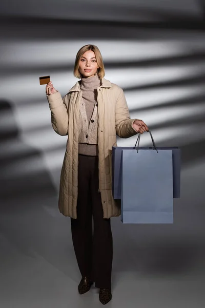 若いです女性で冬のジャケット保持クレジットカードとショッピングバッグ上の抽象グレーの背景 — ストック写真