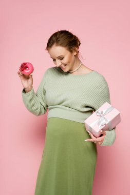 Yeşil kıyafetli mutlu hamile kadın pembe arka planda göbeğine bakarken elinde donut ve hediye tutuyor. 