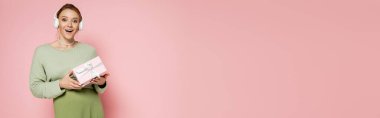 Heyecanlı kulaklıklı hamile kadın pembe arka planda hediyesini tutuyor. 