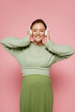 Cheerful pregnant woman in sweater listening music in headphones on pink background  clipart