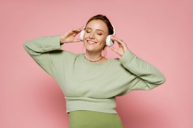 Pozitif hamile kadın pembe arka planda kulaklıkla müzik dinlerken gözlerini kapatıyor. 