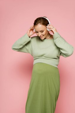 Yeşil kazaklı mutlu hamile kadın kulaklık takıyor ve pembe arka planda göbeğine bakıyor. 