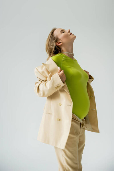 Stylish pregnant woman touching beige jacket while standing isolated on grey 