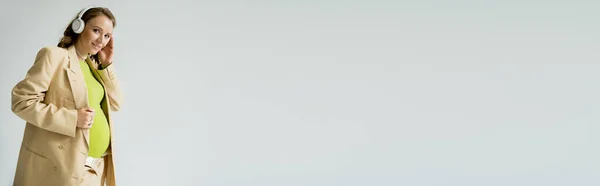 Вагітна Жінка Стильній Куртці Навушники Дивиться Камеру Ізольовано Сірому Банер — стокове фото