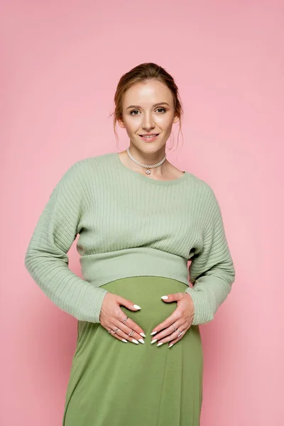 穿着绿色衣服 看着用粉色隔离的相机的正怀孕妇女的画像 — 图库照片