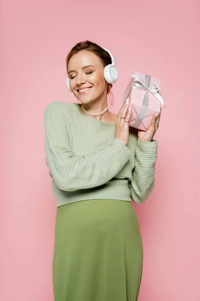 戴着耳机的快乐孕妇拿着礼物 带着用粉色隔离的蝴蝶结 — 图库照片