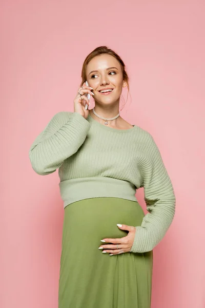 穿着绿色衣服的孕妇笑着用智能手机与粉色隔离 — 图库照片