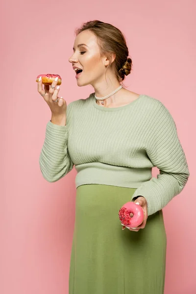 穿着绿色毛衣的孕妇拿着用粉色分离出来的甜甜圈 — 图库照片