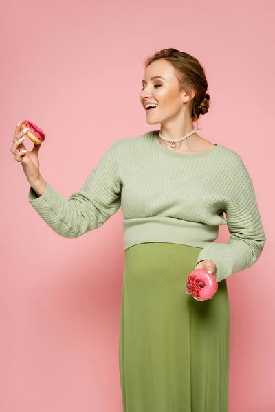 Весела Молода Жінка Зеленому Вбранні Дивиться Пончик Рожевому Фоні — стокове фото