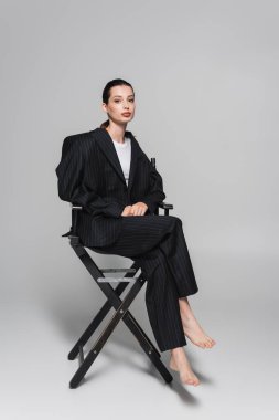 Full length of barefoot woman in striped suit sitting on folding chair on grey background  clipart