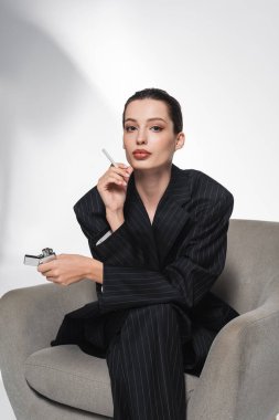 Soyut gri arka planda koltukta otururken elinde sigara ve çakmak tutan çizgili takım elbiseli moda bir kadın. 