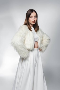 Beyaz elbiseli ve sahte kürk ceketli moda bir kadın soyut gri arka planda kalçaların üzerinde el ele tutuşuyor. 