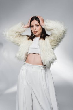 Sahte kürk ceketli, beyaz bluzlu, soyut gri arka planda saçlarına dokunan şık bir kadın. 
