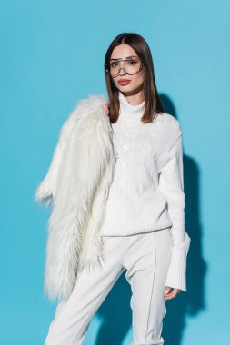 Moda gözlüklü şık bir kadın ve beyaz balıkçı yaka, mavi üzerine beyaz kürk ceket giyiyor.