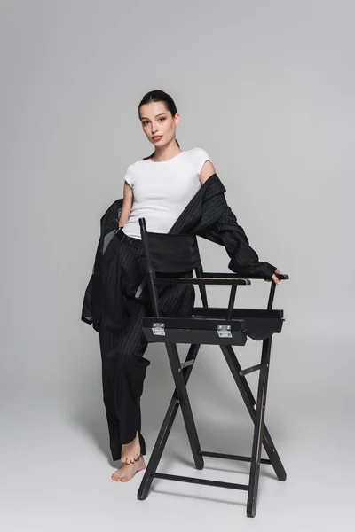 Stylish barefoot woman in black suit and t-shirt posing near folding chair on grey background