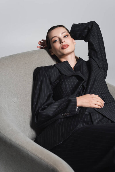 Stylish woman in black suit posing on armchair isolated on grey 