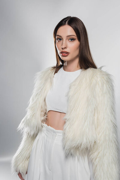 Stylish woman in white faux fur jacket looking at camera on grey background 