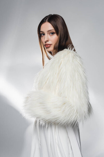 Stylish brunette woman in faux fur jacket looking at camera on abstract grey background 