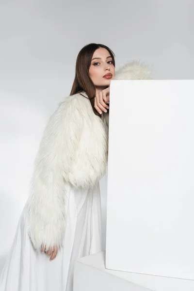 stock image pretty young woman in white faux fur jacket leaning on cube while posing on grey