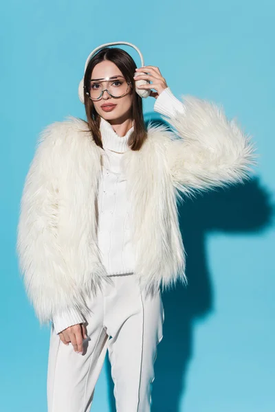 流行の眼鏡と青い上の冬の耳フを調整フェイクファーのジャケットのスタイリッシュな若い女性 — ストック写真