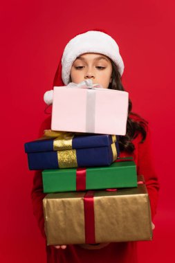 Noel Baba şapkalı ve kazaklı bir kız elinde kırmızı üzerine izole edilmiş bir sürü Noel hediyesi tutuyor.