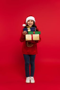 Noel Baba şapkalı ve süveterli mutlu bir kız sürüsü kırmızıda bir sürü Noel hediyesi tutuyor.