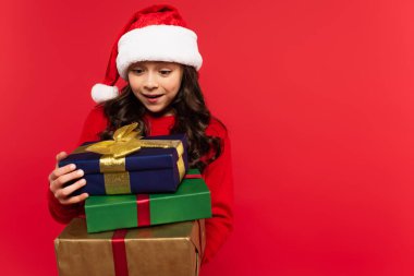 happy girl in santa hat and sweater holding bunch of Christmas presents isolated on red clipart