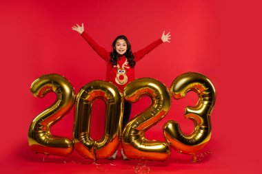 full length of excited girl in sweater standing near balloons with 2023 numbers on red clipart